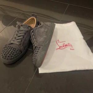 Grå sneakers med nitar från Christian Louboutin - Snygga grå sneakers från Christian Louboutin med nitar på tån och den ikoniska röda sulan. Skorna har snörning och är tillverkade i mocka, vilket ger en lyxig känsla. Perfekta för att ge din outfit en edgy touch.