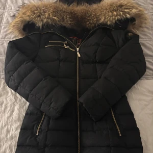 Svart dunjacka från Hollies - Säljer en svart dunjacka från Hollies med pälskantad huva och leopardmönstrat foder. Jackan har dragkedja och flera praktiska fickor. Perfekt för kalla dagar! Observera att den är lite sliten vid kanten på luvan.