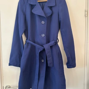 Blå trenchcoat från Vero Moda - Säljer en stilren blå trenchcoat från Vero Moda. Jackan har långa ärmar och knäpps med stora knappar framtill. Den har även ett matchande bälte i midjan för en snygg siluett. Perfekt för vår och höst!