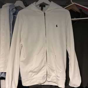 Vit hoodie från Polo Ralph Lauren - Säljer en stilren vit hoodie från Polo Ralph Lauren. Den har en dragkedja framtill och en klassisk logga på bröstet. Perfekt för en avslappnad look. 🏷️