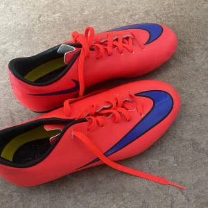 fotbollsskor från Nike - Snygga röda fotbollsskor från Nike med blå och svarta detaljer. Skorna har en slimmad design med snörning för optimal passform. Perfekta för fotbollsplanen!