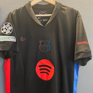 Svart fotbollströja från Nike - Snygg svart fotbollströja från Nike med röda och blå detaljer. Tröjan har korta ärmar och ett stort rött logotryck på framsidan. På baksidan finns ett namn och nummer i rött. Perfekt för fotbollsfans som vill sticka ut!Helt ny med tagg i storlek M en replika