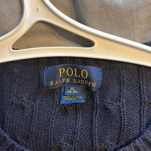Mörkblå stickad tröja från Polo Ralph Lauren - Säljer en snygg mörkblå stickad tröja från Polo Ralph Lauren. Tröjan har ett diskret kabelstickat mönster och en liten röd logotyp på bröstet. Perfekt för kyliga dagar med sin klassiska design och ribbade muddar vid ärmslut och nederkant. Xl men passar till L och M
