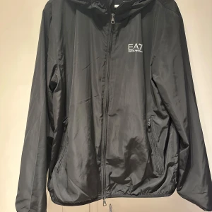 Svart vindjacka från Emporio Armani - Snygg svart vindjacka från Emporio Armani med dragkedja och huva. Jackan har en stilren design med EA7-logga på bröstet och praktiska fickor med dragkedjor. Perfekt för en sportig look.