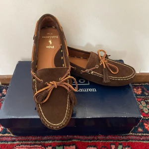  Loafers från Ralph Lauren - Snygga bruna loafers i läder från Ralph Lauren i mocka med dekorativa tofsar och kontrasterande sömmar. Perfekta för en stilren somrig look. Skorna har en klassisk design med rund tå och är lätta att ta på och av. Aldrig använda utomhus endast testat dem inomhus. Köpta i en av ralphlaurens officiella outletbutiker därav priset på lådan. Annars är normalpriset ca 2300. 
