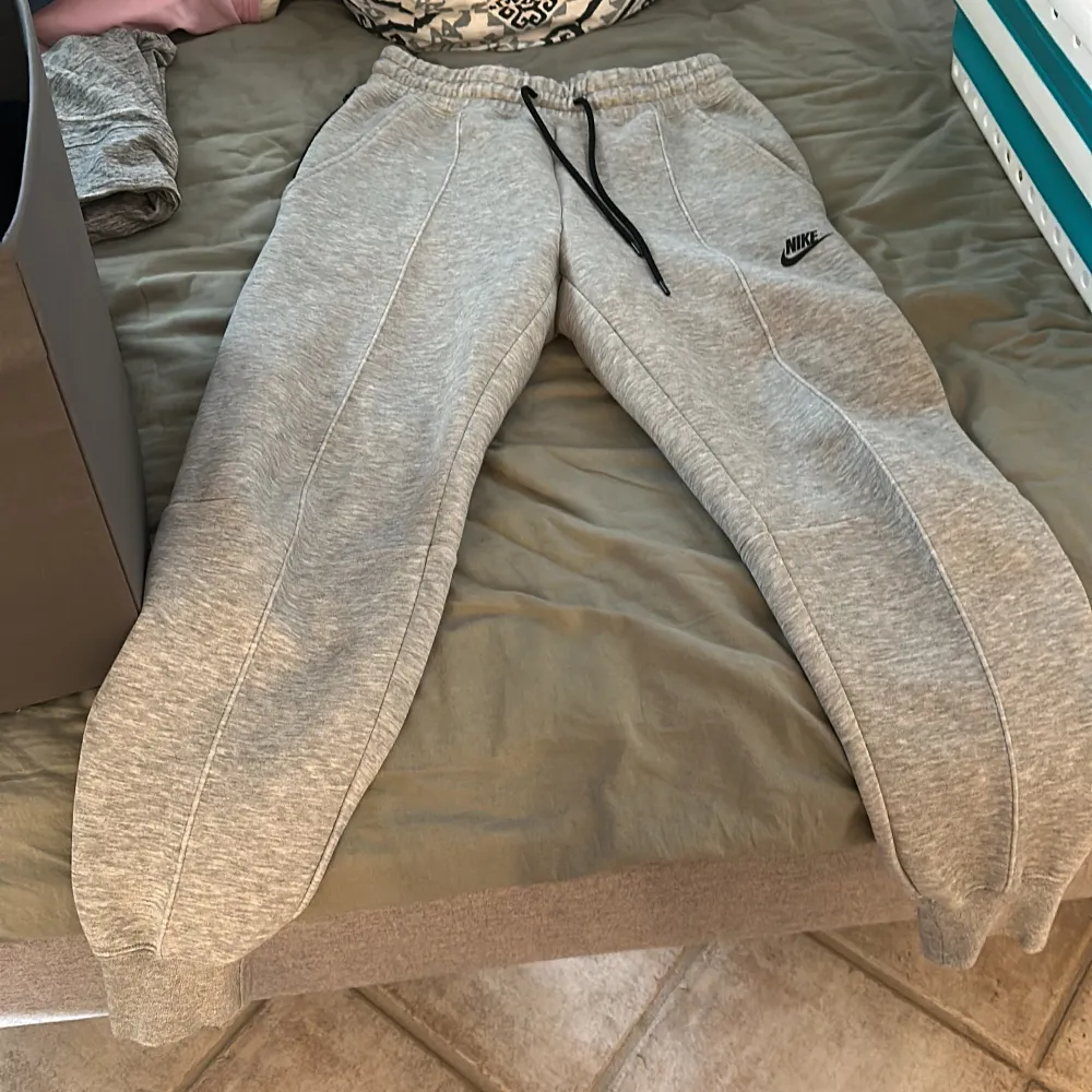 Säljer ett par grå mjukisbyxor från Nike med elastisk midja och svarta snören. Byxorna har praktiska fickor med dragkedja på ena sidan och en broderad Nike-logga på benet. Perfekta för en avslappnad stil. Hör av er🩶. Farkut & Housut.