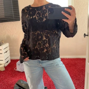 Svart spetsblus med öppen rygg - Snygg svart spetsblus med långärmad design och öppen rygg. Blusen har ett elegant blommönster och knytning i nacken. 
