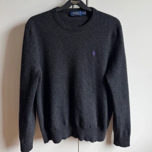 Mörkgrå tröja från Polo Ralph Lauren - Säljer en stilren mörkgrå tröja från Polo Ralph Lauren med lila logga på bröstet. Tröjan har långa ärmar och ribbade muddar vid ärmslut och nederkant. Riktigt skön och snygg men är tyvärr lite för liten för mig. Storlek M