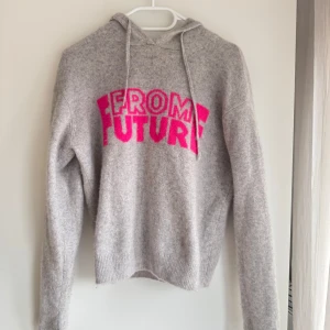 Grå hoodie från From Future - Säljer en supermjuk grå hoodie från From Future i 100% kashmir. Den har en cool rosa text på framsidan och en klassisk huva med dragsko. Perfekt för att hålla sig varm och stilren.