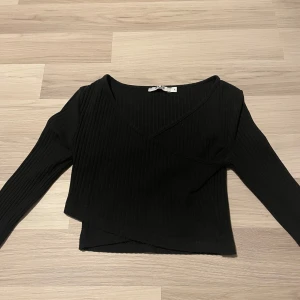 Svart ribbad omlottopp - Snygg svart ribbad omlottopp med långa ärmar. Perfekt för en stilren look. Toppen har en croppad passform och en v-ringad design.