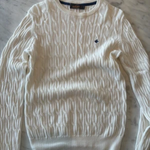 Vit stickad tröja från Morris - Säljer en stilren vit stickad tröja från Morris med ett märke på bröstet. Tröjan har ett klassiskt flätmönster och långa ärmar, den är i nyskick.