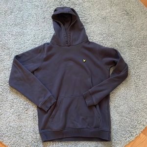 Grå hoodie från Lyle & Scott - Säljer en stilren mörkblå hoodie från Lyle & Scott med en gul broderad logotyp på bröstet. Tröjan har en klassisk känguruficka och en bekväm huva. Perfekt för en avslappnad look!