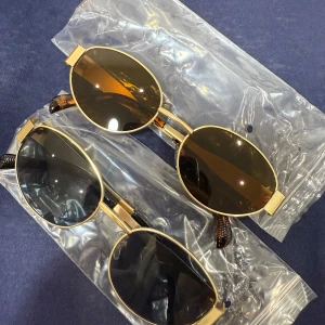 Guldiga solglasögon med ovala linser - Två par stilrena solglasögon med guldiga bågar och ovala linser. Ena paret har mörkbruna linser medan det andra har svarta linser. Perfekta för en solig dag och ger en elegant touch till din outfit.