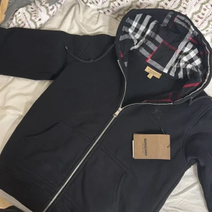 Svart hoodie från Burberry - Säljer en stilren svart hoodie från Burberry med dragkedja och klassiskt rutigt mönster i huvan. Perfekt för en casual look med en touch av lyx. Huvan har ett snyggt mönster i rött, vitt och grått.