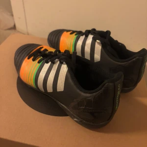 Adidas fotbollsskor med färgglada detaljer - Snygga Adidas fotbollsskor i svart med iögonfallande orange och gröna detaljer. Skorna har klassiska vita ränder och snörning för en säker passform. Perfekta för fotbollsplanen!