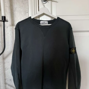 Svart tröja från Stone Island - Säljer en stilren svart tröja från Stone Island med deras ikoniska märke på ärmen. Tröjan har en klassisk rund hals och långa ärmar. Perfekt för en avslappnad och trendig look.