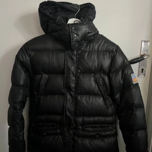 Svart dunjacka från Svea - Säljer en svart dunjacka från Svea med en cool patch på ärmen. Jackan har en pufferdesign och är perfekt för kalla dagar. Den har en hög krage och dragkedja framtill för extra värme. Ett måste för vårens kyligare dagar! 🖤