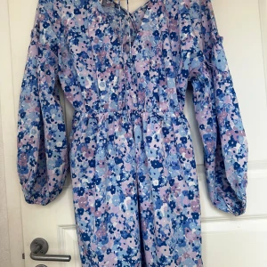 Blommig långärmad klänning från Kappahl - Säljer en söt långärmad klänning från Kappahl med blommigt mönster i blått, lila och rosa toner. Klänningen har v-ringning med knytband och ballongärmar. Perfekt för vår och sommar!