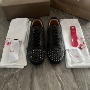 Svarta sneakers med nitar från Christian Louboutin - Säljer ett par svarta sneakers från Christian Louboutin med silverfärgade nitar på tån och klassisk röd sula. Skorna har snörning och är tillverkade i skinn. Perfekta för dig som vill sticka ut med en lyxig och edgy stil.