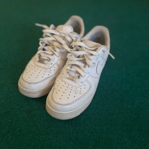 Vita Nike Air Force 1 sneakers - Klassiska vita Nike Air Force 1 sneakers med snörning och rund tå. Skorna har en platt sula och är tillverkade i syntetmaterial med diskreta detaljer. Perfekta för en stilren och sportig look. Mycket fint skick, smart använt!
