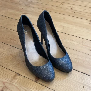 Glittriga pumps - Snygga glittriga pumps i en mörkblå nyans. Skorna har en spetsig tå och hög klack, vilket ger en elegant look. Perfekta för en kväll ute eller en speciell tillställning.