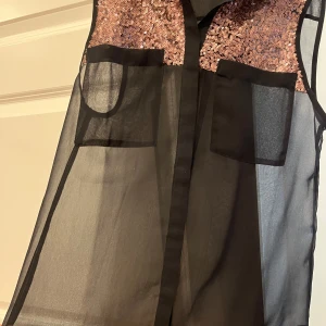 Svart transparent blus med paljetter från Vila - Snygg svart transparent blus från Vila med glittriga paljetter i brons på axlarna. Blusen har en klassisk krage och två bröstfickor. Perfekt för en kväll ute eller en festlig tillställning. ✨