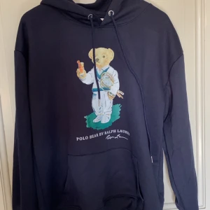 Blå Polo Bear hoodie  - Säljer en snygg mörkblå hoodie från Ralph Lauren med Polo Bear på framsidan. Tröjan har en klassisk känguruficka och justerbar huva med snören. Perfekt för en avslappnad och trendig stil!