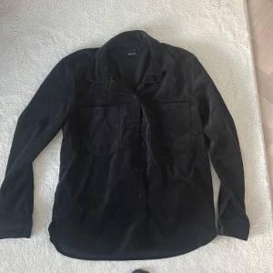 Svart manchester skjorta från Gina Tricot - Snygg svart manchester skjorta från Gina Tricot med långa ärmar och knappar framtill. Skjortan har två stora bröstfickor och en klassisk krage. Perfekt för en stilren look.