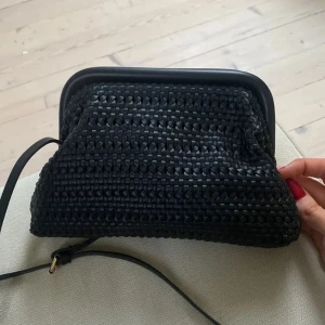 Svart väska med flätad design - Snygg svart väska med en unik flätad design. Väskan har en justerbar axelrem och en praktisk öppning med metallram. Perfekt för att bära dina nödvändigheter med stil.