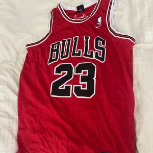 Chicago Bulls baskettröja Jordan 23 - Röd Chicago Bulls baskettröja med nummer 23 och namnet Jordan på ryggen. Tröjan har svarta och vita detaljer runt ärmhål och hals, samt stora tryck fram och bak. Perfekt för basketfans! Och är Storlek L