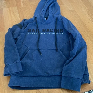 Blå hoodie från Sail Racing, stl S herr - Mörkblå hoodie från Sail Racing med trycket 'SAIL RACING ANTARCTICA EXPEDITION' på bröstet. Tröjan har huva med dragsko och en stor magficka. Herr storlek S. 