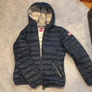 Mörkblå dunjacka med huva från Colmar - Säljer en mörkblå dunjacka från Colmar med huva och dragkedja. Jackan har två snedställda fickor med dragkedja och quiltad design. Insidan är ljusgrå och jackan har en sportig look, perfekt för kyliga vår dagar. OBS!!! En liten defekt på sista bilden som är fixat och syns knappt.