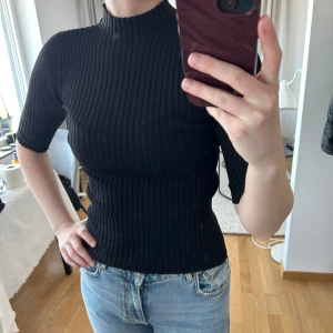 Svart ribbad topp med halv polo - Säljer en svart ribbad topp med halv polokrage och korta ärmar. Tröjan är tight i modellen och har en stretchig passform som sitter snyggt på kroppen. Perfekt att styla med jeans eller kjol för en clean look. Använd 1 gång.