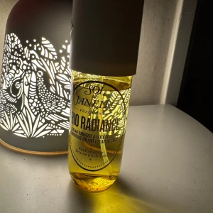 Parfym Sol de Janeiro Rio Radiance - Fräsch och exotisk parfym från Sol de Janeiro, Rio Radiance. Flaskan är genomskinlig med en vit kork och innehåller en gyllene vätska. Doften har inslag av solig tuberos och kokosmjölk, perfekt för en tropisk känsla.