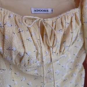 Gul blommig klänning från ADOORE - Charmig gul klänning från ADOORE med blommigt mönster och puffiga kortärmar. Klänningen har en knytning framtill och rynkade detaljer längs sidorna för en smickrande passform. Perfekt för soliga dagar!