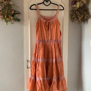 Korall klänning med spetsdetaljer - Charmig korallfärgad klänning med knytning framtill. Klänningen har breda axelband och volangkant nedtill, vilket ger en bohemisk känsla. Perfekt för varma sommardagar.