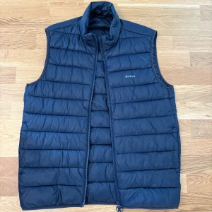 Marinblå dunväst från Barbour - Säljer en marinblå dunväst från Barbour med ståkrage och dragkedja framtill. Västen har ett quiltat mönster och är perfekt för lager-på-lager. Klassisk och stilren design med Barbour-logga på bröstet. Nypris 1899kr