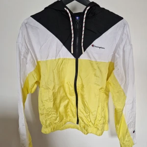 Gul och vit vindjacka från Champion - Säljer en snygg vindjacka från Champion i färgerna gul, vit och svart. Jackan har huva med dragsko, dragkedja framtill och elastisk nederkant. Perfekt för vår och sommar när du vill ha en lätt jacka med cool retrostil. Använd ett fåtal gånger så i mycket bra skick!🤗