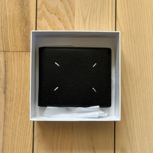 Maison Margiela Plånbok - Säljer en stilren svart plånbok från Maison Margiela i skinn. Plånboken har flera kortfack och klassisk design med de ikoniska vita sömmarna på framsidan. Perfekt för dig som gillar minimalistisk och lyxig stil. Kan sänka pris vid snabb affär!