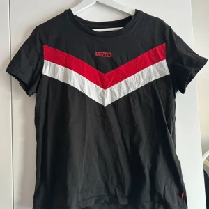 Svart t-shirt från Levi's med röd och vit detalj - Svart t-shirt från Levi's med ett snyggt V-format mönster i rött och vitt över bröstet. Klassisk passform och korta ärmar. Tillverkad i mjuk bomull och har Levi's-logga framtill.