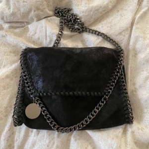 Svart axelväska med kedjedetaljer - Säljer en svart axelväska med grov silverfärgad kedja som axelrem och runt kanterna. Väskan har ett lock med magnetknäppning och insidan är fodrad med mönstrat tyg. Perfekt för att lyfta vilken outfit som helst. Gör allt till 10/10