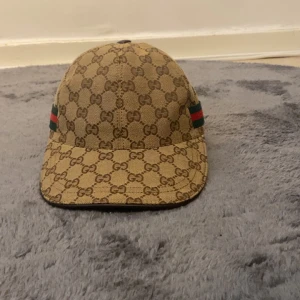 Beige keps från Gucci med GG-mönster - Säljer en beige keps från Gucci med klassiskt GG-monogram över hela kepsen. Kepsen har gröna och röda ränder på sidorna och justerbart spänne bak. Perfekt för dig som gillar lyxiga accessoarer och streetstyle.