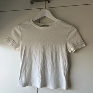 Vit topp från Zara - Säljer en vit smått ribbad topp från Zara. Toppen har korta ärmar med pärldetaljer och en rund halsringning. 