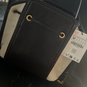 Brun och vit axelväska från Zara - Snygg brun och vit axelväska från Zara med kontrasterande detaljer. Väskan har en praktisk dragkedja och dekorativa metallringar. Perfekt för att bära dina dagliga nödvändigheter med stil.