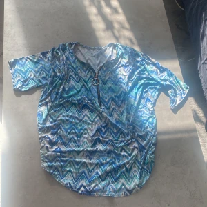 Mönstrad t-shirt med dragkedja - Säljer en färgglad t-shirt med ett zigzag-mönster i blå, grön och vit. T-shirten har korta ärmar och en dragkedja framtill med en rund detalj. Perfekt för en avslappnad stil.