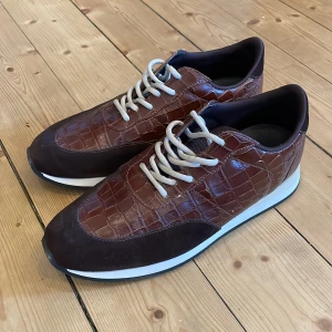 Bruna sneakers - Snygga bruna sneakers från Giuseppe Zanotti med krokodilmönstrad yta och vita snören. Skorna har en rund tå och en kontrasterande vit sula. Vid snabb affär går priset att diskutera👍