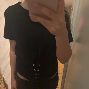 Svart kortärmad topp med snörning - Säljer en svart kortärmad topp med snörning framtill. Toppen har en tight passform och är perfekt för en stilren look.