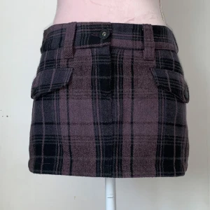 Rutig kortkjol med fickor - Snygg rutig kortkjol från i lila och svart. Kjolen har praktiska fickor fram och en knappdetalj i midjan. Ullblandning. 