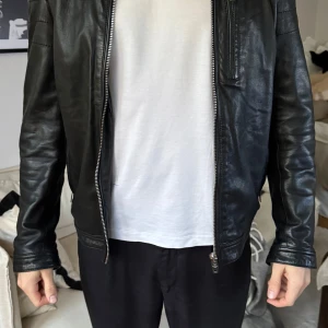 Svart skinnjacka med dragkedja - Snygg svart skinnjacka med dragkedja framtill och en bröstficka med dragkedja. Jackan har en stilren design med långa ärmar och knappar vid kragen.