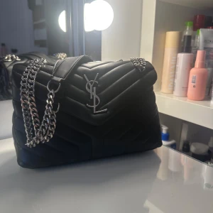 Svart quiltad axelväska från Saint Laurent - Snygg svart axelväska från Saint Laurent med quiltad design och silverfärgade kedjedetaljer. Väskan har ett stort YSL-emblem på framsidan och en praktisk dragkedja inuti. Perfekt för att ge en lyxig touch till din outfit.